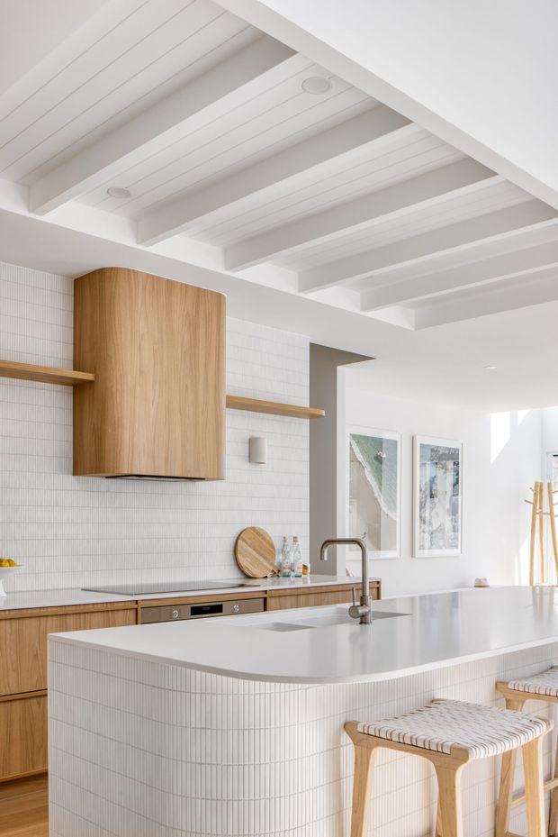 Bright, refined kitchen with a large white island, integrated sink and brushed metal tap, light timber cabinetry, and a curved timber rangehood enclosure. The walls and island are clad in fine white vertical-line tiles, while the ceiling features exposed white-painted beams and panelled infill. Timber stools, open shelving, understated wall lights, and framed artwork complete the airy interior.