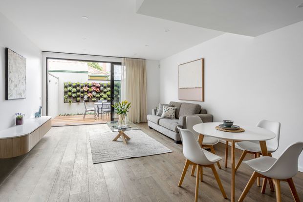 Bright, contemporary open-plan living and dining area with pale timber flooring, a neutral grey sofa, white dining table and sculptural white chairs. Full-height sliding glass doors open to a small planted outdoor courtyard, while sheer curtains, restrained artwork, built-in media cabinetry and a light textured rug create an airy, refined interior.