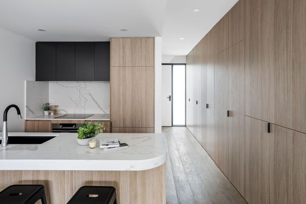A bright contemporary kitchen with a large curved island, integrated black appliances and sink, pale veined stone benchtops and splashback, and extensive light timber cabinetry. A long timber-lined wall and hallway lead toward a glazed entry door, while black stools and dark upper cabinets provide contrast.