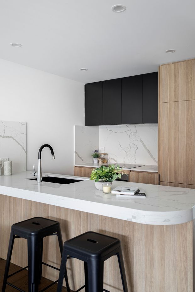 A contemporary kitchen with a large island featuring a white veined stone benchtop, integrated sink and black gooseneck tap. The island has light timber-grain cabinetry and two black metal stools. Matching light timber cabinetry runs along the right wall, contrasted with tall matte-black overhead cabinets and a white marble-look splashback. The room has white walls and ceiling with recessed downlights, creating a clean, refined, neutral interior.