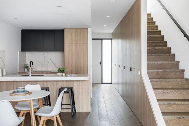 Bright contemporary open-plan kitchen and dining area opening onto a hallway and staircase. The kitchen features light timber cabinetry, dark overhead cupboards, a pale stone-look splashback and benchtop, and a compact island with seating. A small round dining table with modern chairs sits in the foreground. The hallway continues beside full-height timber wall panelling and a timber staircase with a simple dark handrail.