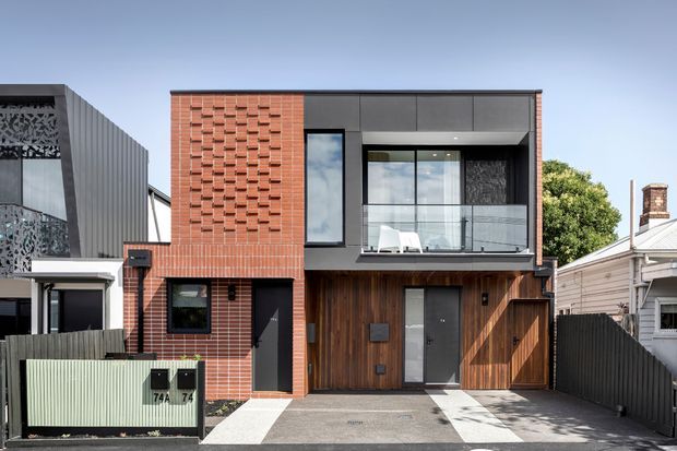 Street-facing view of a narrow contemporary multi-storey townhouse or residence. The facade combines red brickwork with a decorative projecting brick pattern, dark metal panels and framing, vertical timber-look cladding around the garage and entry, and a glazed upper-level balcony. Adjacent urban residential buildings, fencing, driveway paving, and mature trees are visible.