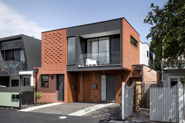 Street-facing view of a contemporary multi-level urban residence with a flat roof, red brick upper and lower facade sections, dark charcoal wall panels, extensive upper-level glazing with a glass balustrade, and warm vertical timber cladding around the recessed entry. A paved driveway, timber fencing, and neighbouring homes are visible.