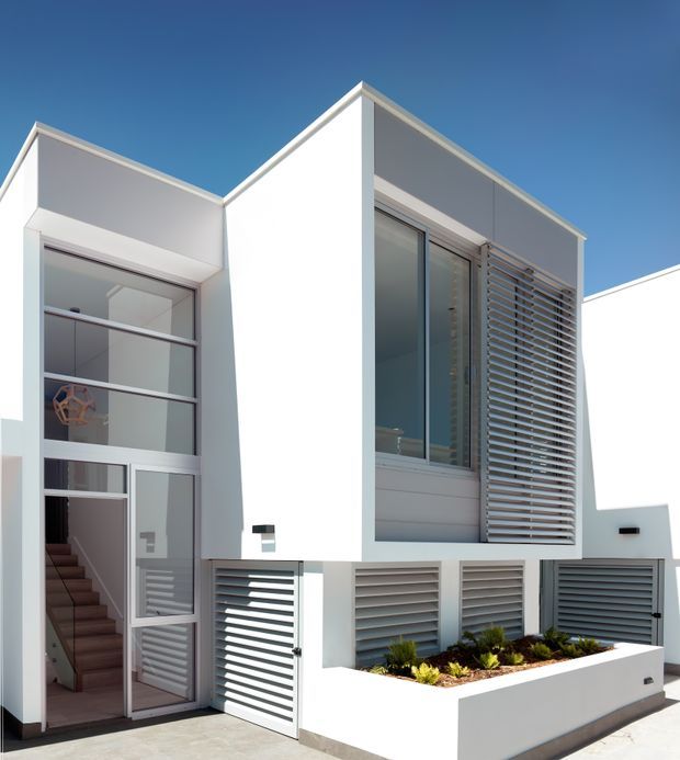 Exterior view of a contemporary multi-level residence with crisp white geometric volumes, steeply sloped roof planes, extensive glazing, and horizontal louvre screens over windows and garage openings. A recessed glazed entry reveals an internal staircase, while a raised white planter with low landscaping fronts the facade.