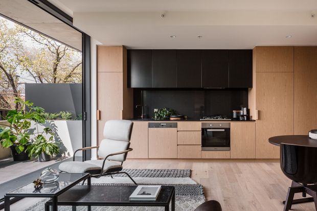 Contemporary open-plan kitchen and living area with a full-height glazed wall, pale timber cabinetry, dark upper kitchen joinery and splashback, integrated stainless-steel appliances, and light timber flooring. A lounge chair, coffee table, rug and grand piano occupy the living zone, with leafy planting visible beyond the glazing.