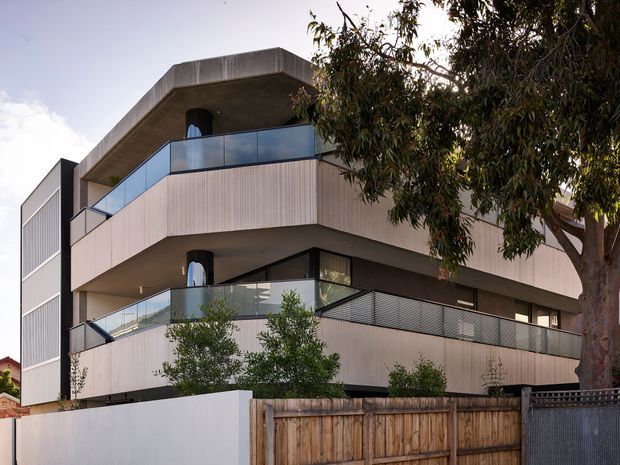 Exterior view of a contemporary multi-storey residential building with broad projecting balconies, pale vertically ribbed facade panels, dark recessed soffits, glass balustrades, and a flat concrete roof slab. Mature trees and timber boundary fencing partially screen the lower level.