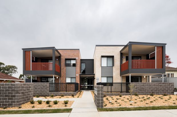 Street-facing view of a contemporary multi-unit residential development with two matching two-storey dwellings. The buildings feature light brick walls, dark charcoal framing and cladding, recessed upper-level balconies with red vertical privacy screens, glazed entry doors, and low dark masonry boundary walls. Sparse landscaped front gardens and a central pedestrian path lead to the shared entrance.