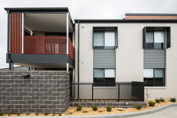Contemporary multi-storey residential facade with a projecting upper-level balcony, vertical red timber screening, dark metal framing and balustrades, pale rendered walls, and charcoal brick masonry boundary walls. Windows feature dark frames and grey horizontal privacy panels, with a narrow landscaped setback and paved street edge.