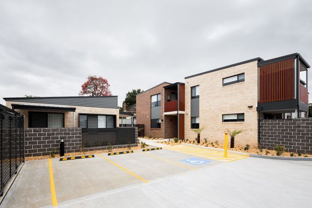 Exterior view of a contemporary multi-unit residential development with several low-rise buildings arranged around a paved parking area. The buildings feature pale brick walls, dark metal roof edges and frames, black masonry boundary walls, and prominent vertical timber or rust-toned screening on upper balconies. Accessible parking markings and yellow kerb lines are visible in the foreground.