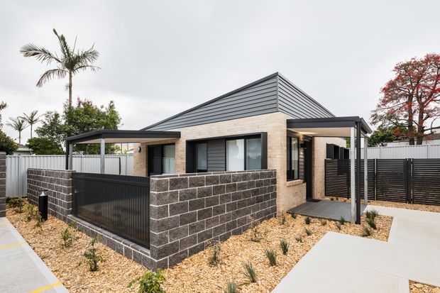 Single-storey contemporary home exterior with a distinctive folded gable roof, dark horizontal cladding, pale brick walls, black-framed glazing, covered entry and a private front courtyard enclosed by dark masonry and slatted fencing. The landscaped frontage uses light gravel and sparse low planting, with a concrete path and driveway.