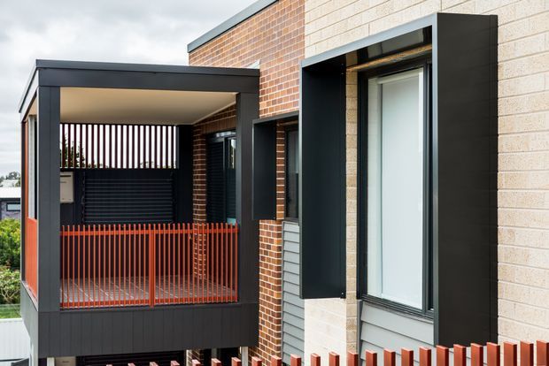 Exterior architectural detail of a contemporary multi-level residential building, showing a projecting covered balcony with dark cladding, vertical red-orange balustrades, brickwork, pale masonry walls, and large black-framed windows.