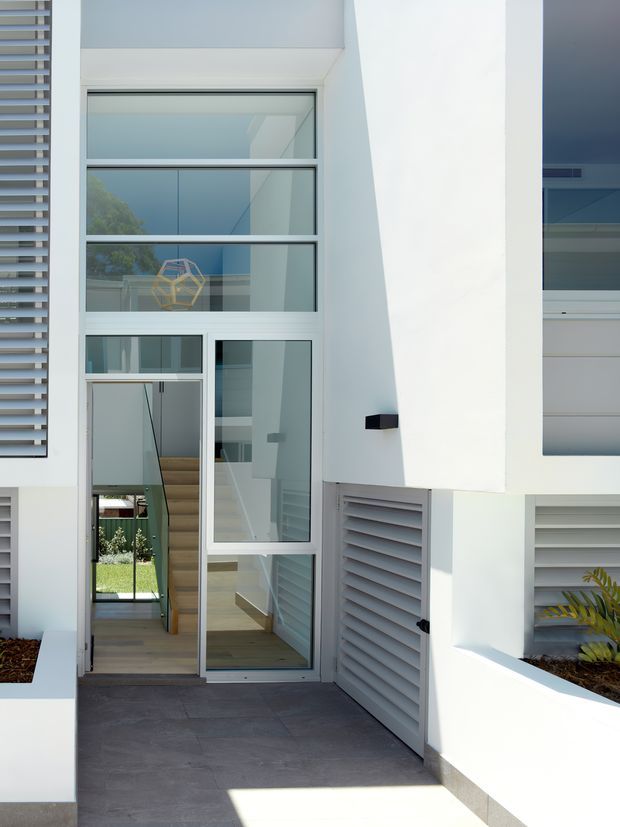 Contemporary white multi-level residential facade viewed from the entry path, featuring a tall glazed entry with stacked horizontal window panels, a glazed front door, grey louvred screens, crisp geometric wall planes, and dark tiled paving. Interior stairs and a small landscaped courtyard are visible through the glazing.