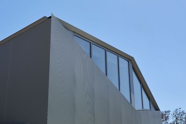 Exterior architectural detail of a contemporary multi-storey building with a sharply angular facade, vertically ribbed grey cladding, and a prominent band of tall glazed windows beneath a sloping roofline. The image is photographed from below against a clear blue sky.