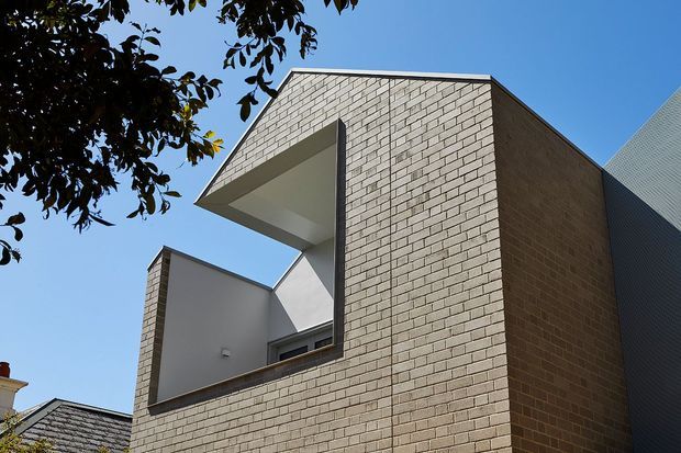 Contemporary urban building exterior photographed from below, featuring a boxy form clad in pale textured brick, a deep recessed balcony or window opening, sharp projecting roof or canopy planes, and a contrasting dark corrugated side wall. Mature tree foliage frames the upper left against a clear blue sky.