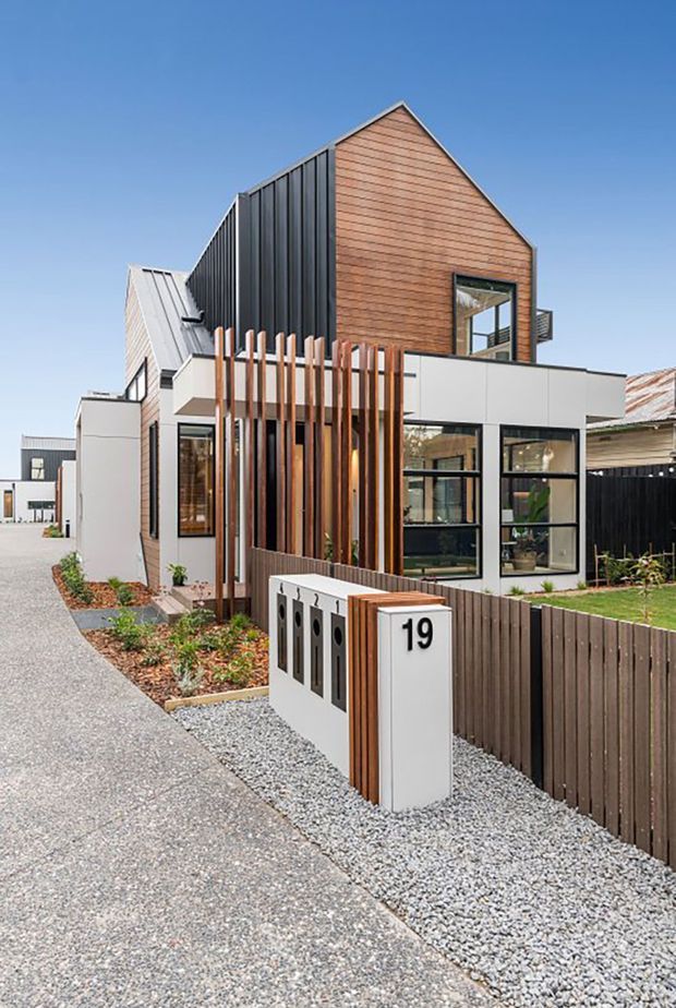 A contemporary multi-level townhouse facade viewed from the street, featuring a steep gabled form clad in warm horizontal timber boards, dark vertical metal cladding, white rendered or panelled lower volumes, and large black-framed windows. Vertical timber screening and a modern multi-mailbox entry structure sit beside a gravel-paved frontage and timber fence. Adjacent attached or closely spaced homes are visible in the background under a clear blue sky.