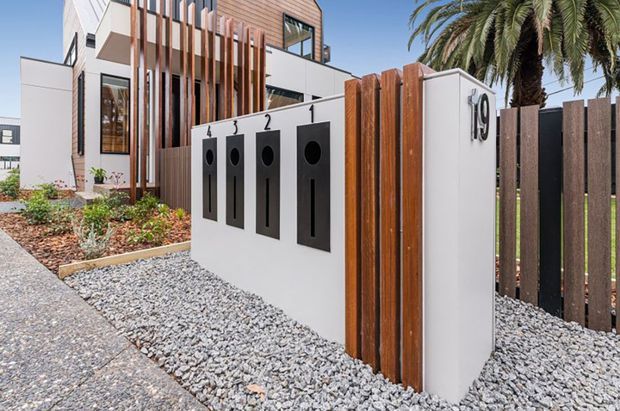 Contemporary multi-storey residential facade and street entry, featuring a white rendered boundary wall with vertically mounted dark letterbox panels, timber screening battens, a dark vertical-slat fence, and a gravel forecourt. The building behind combines white rendered walls, warm timber cladding, large windows, and strong vertical architectural elements, with tropical palm planting beside the entry.