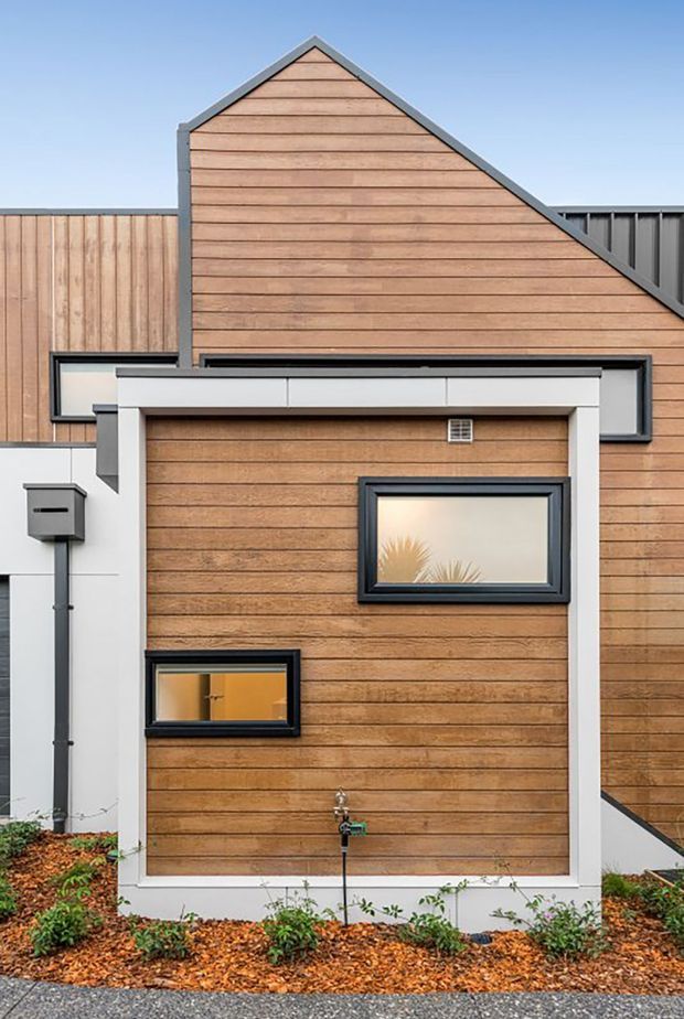 Close-up exterior view of a contemporary residential facade with warm horizontal timber cladding, dark metal-framed windows, crisp white trim, and intersecting gabled roof forms. A small landscaped planting bed with mulch sits at the base, while the composition highlights the modern geometric facade and contrasting materials.