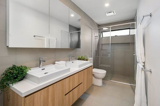 Bright contemporary bathroom with a double vanity, white integrated basins, timber-look floating cabinetry, large mirrored storage, a frameless glass shower, toilet and wall-mounted towel rails. Neutral beige and white finishes create a clean, refined appearance, with small plants adding natural contrast.