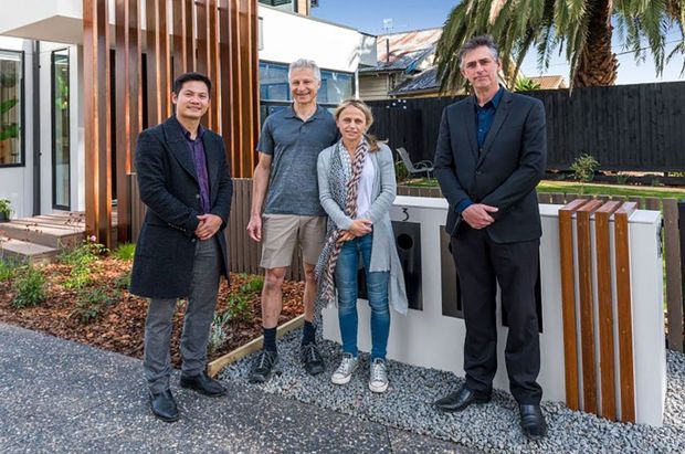 A group of four adults posing outside a contemporary residential property. The home features white rendered walls, large dark-framed glazing, and a prominent vertical timber-clad facade screen. A landscaped entry garden, gravel paving, black boundary fencing, palm trees, and a timber-and-metal front fence are visible.