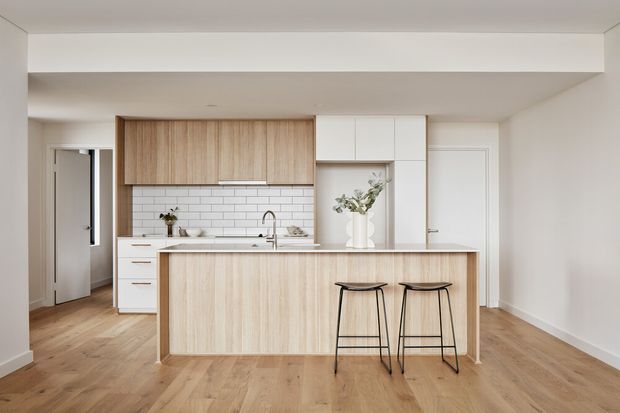 A bright, minimalist contemporary kitchen with a large light-wood island, white cabinetry, white subway-tile splashback, timber flooring, and two black-framed bar stools. The open-plan space includes a concealed or partially visible hallway/entry area and simple white walls and ceiling.