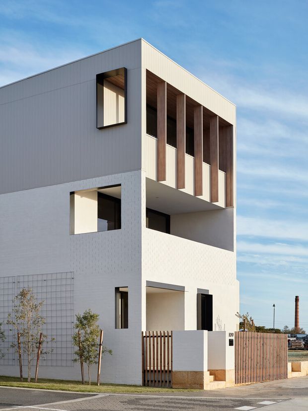 A contemporary multi-storey residential building with a crisp white facade, deep recessed openings, and a prominent upper-level screen of tall vertical timber-lined bays. The design combines rectilinear massing, sheltered balconies, narrow dark-framed windows, and a timber slat front gate. It is photographed externally from the street in clear daylight, with minimal landscaping and an urban context.
