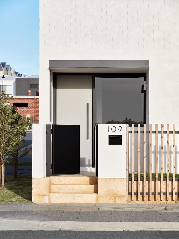 Street-level view of a contemporary residential front entry. A minimalist white rendered facade features a recessed doorway with a pale solid door and narrow vertical pull, a dark glazed sidelight, sandstone steps and plinth, black metal gate, timber-toned vertical slat fencing, and a white mailbox pillar marked 109. Nearby brick and modern townhouse forms are visible in the background.