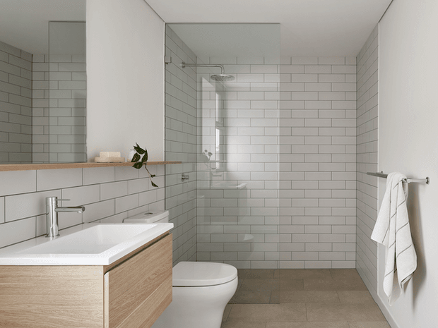 Bright contemporary bathroom with a floating light-wood vanity, white rectangular basin, chrome tapware, wall-to-wall rectangular white tiles, a frameless glass walk-in shower, and a wall-hung toilet. A large mirrored cabinet or wall mirror reflects the tiled room, while a simple towel rail and folded white towels sit on the right wall. The neutral palette and clean linear detailing create a calm, minimal appearance.