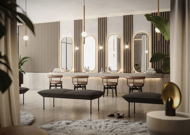 A refined contemporary salon or dressing area with a long stone-topped vanity and multiple individual makeup stations. Tall arched mirrors are set against alternating vertical fluted wall panels and light stone wainscoting. Sculptural pendant lights hang above the vanity, while dark upholstered benches, a textured rug, indoor plants, and minimalist decorative objects add warmth and contrast. The room has a polished, softly lit, luxurious atmosphere.