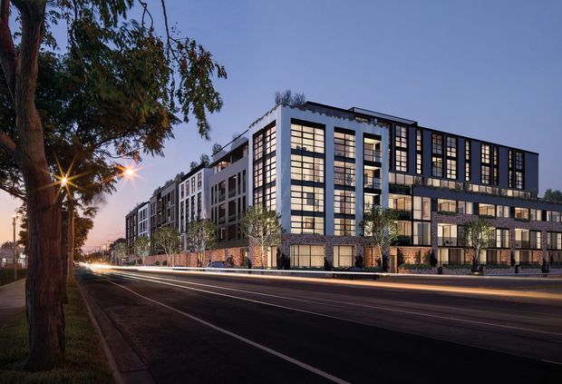 Twilight exterior view of a large contemporary multi-storey urban building lining a broad roadway. The facade combines dark and light vertical panels, extensive gridded floor-to-ceiling windows, projecting balconies, ground-level masonry detailing, and rooftop planting. Street trees, paved footpaths, and illuminated windows emphasize its urban residential or mixed-use character.