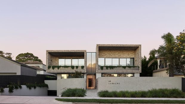 Street-facing view of a contemporary two-storey residential building with two prominent rectangular upper-level volumes, textured concrete or rendered surrounds, extensive glazing, planted balcony edges, and a low pale boundary wall. The central entry is recessed between the two wings, with landscaped grasses and palms around the frontage.