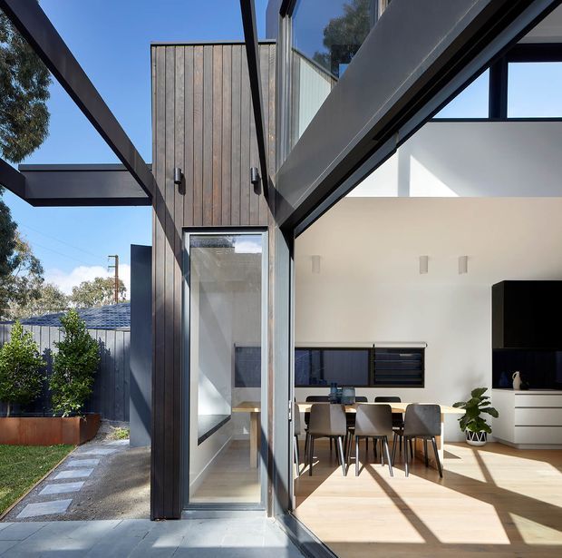 A contemporary residential extension viewed from an outdoor side path, featuring dark vertical timber cladding, expansive black-framed glazing, and strong exposed structural beams. Through the open glazed connection, a bright open-plan dining and living area is visible with a timber dining table, upholstered chairs, white walls, pale timber flooring, and built-in media cabinetry. The composition emphasises indoor–outdoor connection, crisp geometric forms, and abundant natural light.