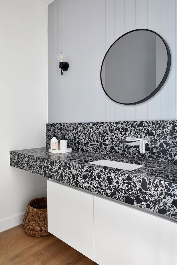 A contemporary bathroom vanity with a wall-mounted white basin and chrome tap, set beneath a large round black-framed mirror. The vanity and matching backsplash feature a striking black-and-white terrazzo surface, with white handleless cabinetry below. Vertical white wall panelling, a black wall sconce, timber flooring, and a woven basket complete the clean, graphic scheme.