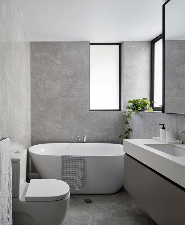 A contemporary grey-tiled bathroom featuring a freestanding white bathtub, wall-mounted vanity with integrated basin, toilet, black-framed frosted windows, and a small trailing plant. The space has a clean monochromatic palette with dark grey cabinetry and contrasting black fixtures and frames.