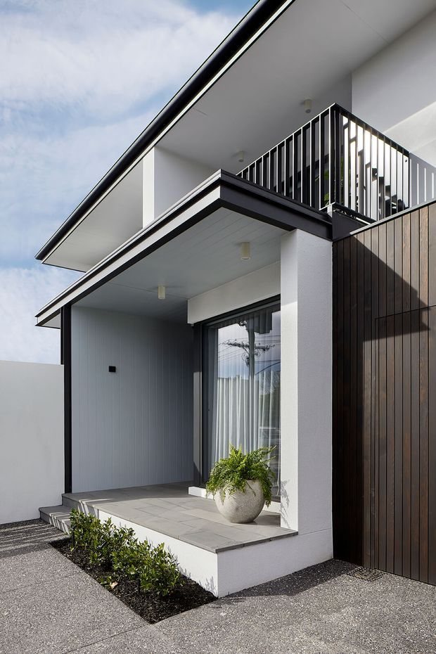 Modern residential front facade viewed from the driveway, featuring crisp white rendered walls, dark vertical timber-look cladding, black metal balcony balustrades, deep roof eaves, and a covered entry porch with grey tiled paving. A glazed entry door, potted fern, and narrow planted garden bed are visible.
