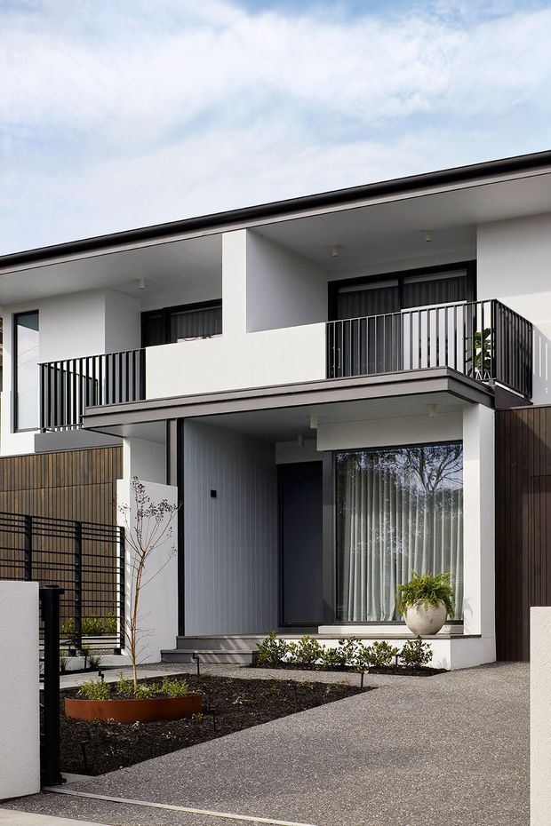 Exterior view of a contemporary two-storey residence with a clean white rendered facade, strong horizontal roof and balcony lines, dark metal balcony railings, large windows, and a recessed ground-floor entry. The home is set behind a paved driveway with minimalist garden beds, black metal fencing, and dark vertical cladding accents.