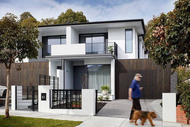 Contemporary two-storey urban residence with a crisp white rendered facade, low-pitched roof, projecting upper balcony, dark vertical timber screening, and black metal fencing. The street-facing frontage includes a gated entry, driveway, landscaped planting, and a partially visible garage or lower-level opening.
