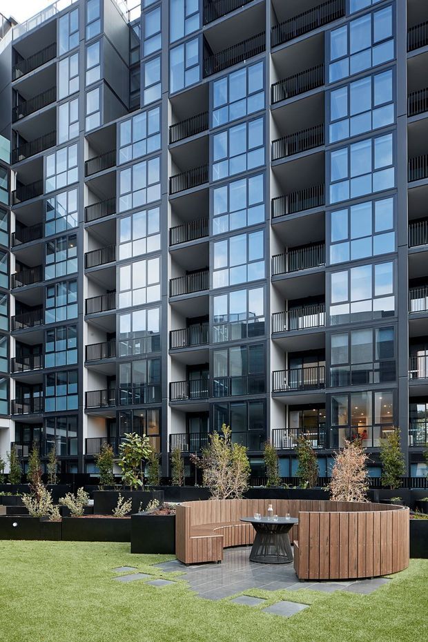 Exterior view of a contemporary multi-storey apartment building with dark metal-framed floor-to-ceiling glazing, recessed balconies, and black balustrades. The foreground contains a landscaped communal courtyard with lawn, raised planters, stone stepping pavers, and curved timber outdoor seating around a small table.