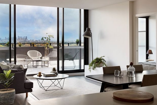 Bright contemporary open-plan living and dining area with floor-to-ceiling black-framed glazing opening to a balcony terrace. The room features a neutral palette, dark timber dining table, upholstered seating, a sculptural coffee table, indoor plants, and a city skyline view beyond the balcony.