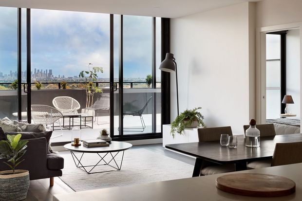 A bright, contemporary open-plan living and dining area with floor-to-ceiling black-framed glazing opening onto a balcony. The room includes a dark sofa, round coffee table, large rectangular dining table, neutral rug, indoor plants, and a compact kitchen bench in the foreground. A city skyline is visible beyond the balcony.