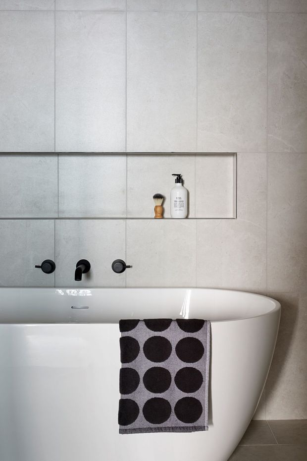 A contemporary bathroom featuring a freestanding white bathtub, wall-mounted matte black tapware, large light grey stone-look wall tiles, and a recessed tiled shelf containing a soap dispenser and shaving brush. A monochrome geometric towel hangs over the bath.