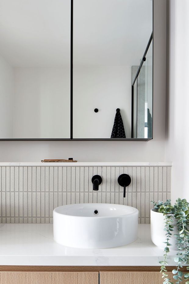 A bright, contemporary bathroom vanity with a white round vessel basin on a white countertop, black wall-mounted tapware, pale vertically grooved wall tiles, light timber cabinetry, and a large black-framed mirrored cabinet. A potted trailing plant adds greenery beside the basin.