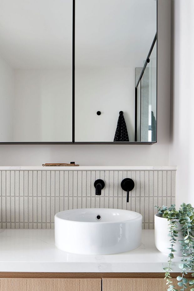 A bright, minimalist bathroom vanity featuring a white vessel basin on a pale benchtop, matte black wall-mounted tapware, and a vertical light-toned tiled splashback. A large black-framed mirrored cabinet spans the wall above, while light timber cabinetry and a trailing potted plant add warmth and greenery.