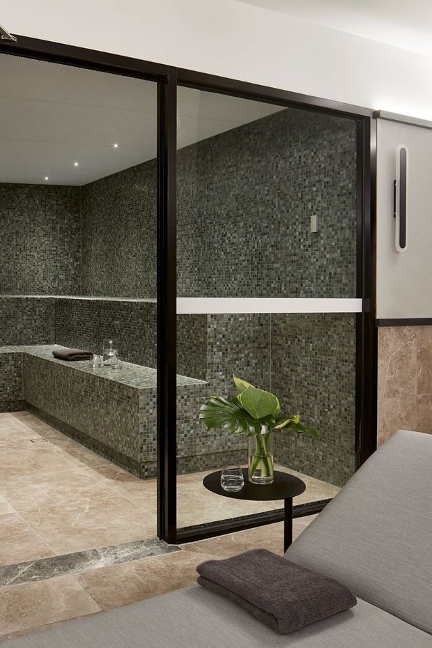 A refined spa-like bathroom viewed from an adjacent lounge or relaxation area. The bathroom is enclosed by dark-framed glass partitions and features full-height small mosaic tile walls in muted grey-green tones, a long tiled bench, recessed ceiling spotlights, and large beige stone-look floor tiles. A grey upholstered daybed or lounge chair, small black side table, and vase of tropical foliage occupy the foreground.
