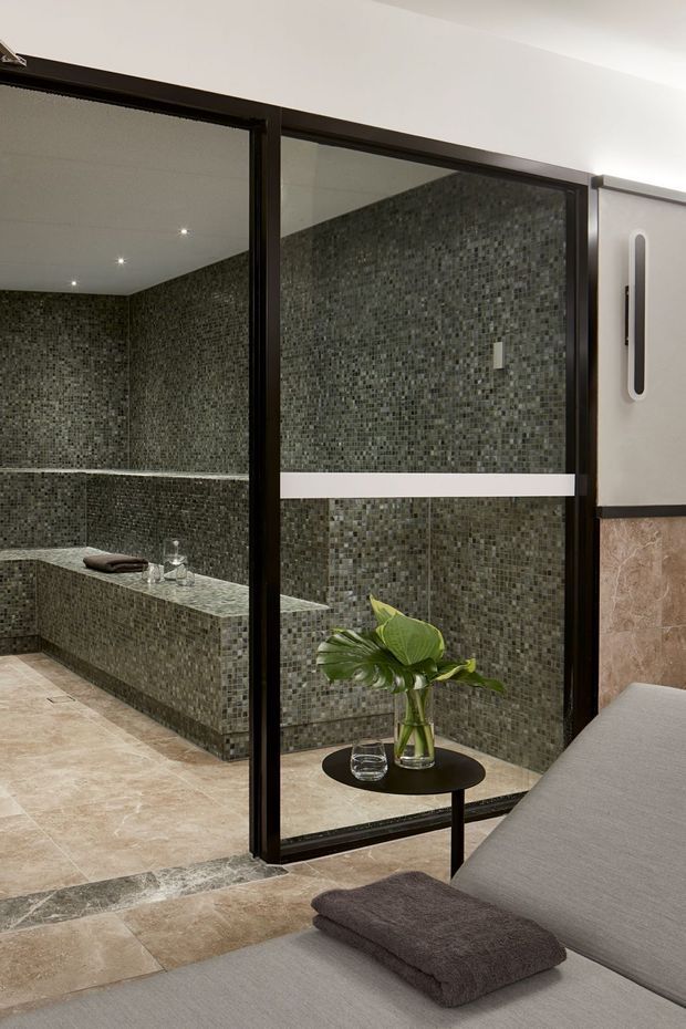 A refined spa-style bathroom viewed through a dark-framed glass door, featuring full-height small-format grey mosaic wall tiling, a long built-in tiled bench, beige stone-look floor tiles, and recessed ceiling lights. In the foreground is a minimalist grey upholstered lounge or treatment bed with folded towels and a small black side table holding a vase of greenery.
