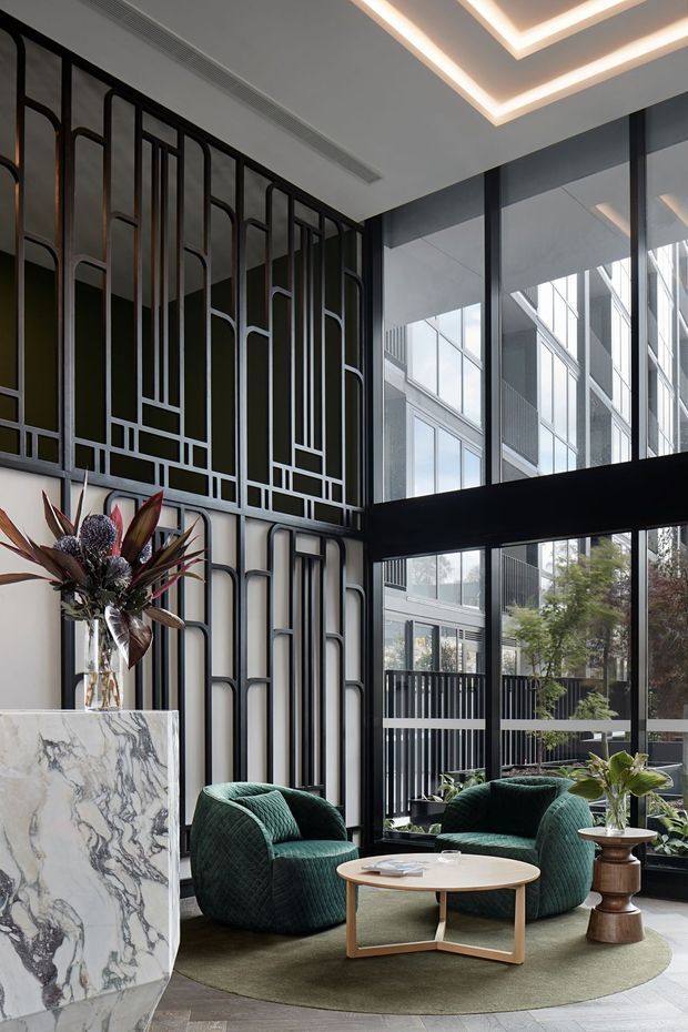 A sophisticated double-height lobby or lounge with dramatic black metal geometric screening, expansive black-framed glazing, layered recessed ceiling lighting, and a large veined marble pedestal. Two deep green sculptural lounge chairs surround a round light timber coffee table on a muted circular rug, with indoor planting and floral arrangements adding organic contrast. The space has a refined, luxurious contemporary feel with strong Art Deco-inspired detailing.