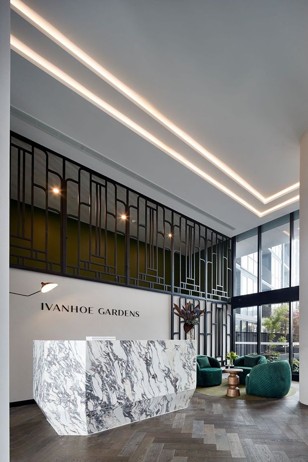 A sophisticated apartment or commercial lobby reception area featuring a large white-and-grey veined stone reception desk, dark herringbone timber flooring, a sculptural black metal screen with geometric curved detailing, high glazing, and a bright contemporary coffered ceiling with recessed linear lighting. Teal lounge chairs and greenery create a refined seating area beside the reception desk. The wall is branded “IVANHOE GARDENS.”