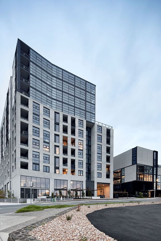 Exterior street-level view of a contemporary multi-storey apartment or mixed-use building. The main tower has a light grey concrete or rendered facade, extensive dark-framed glazed curtain walling at the upper levels, recessed balconies, and ground-floor commercial glazing. A smaller adjoining dark-and-grey modern building is visible to the right, with paved streetscape and landscaped gravel in the foreground.