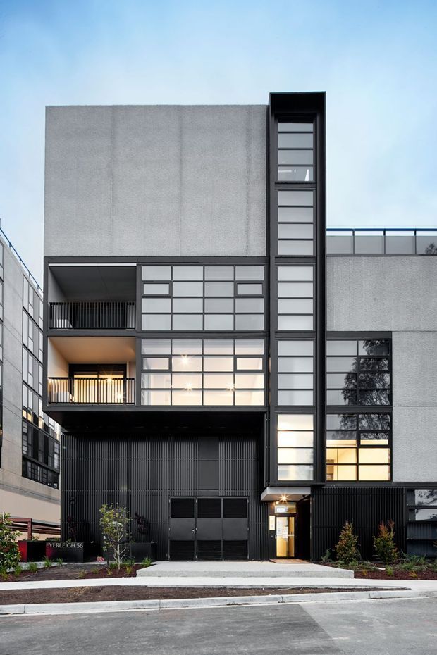 Street-level exterior photograph of a contemporary multi-storey urban residential or mixed-use building. The facade combines large textured grey panels, extensive dark-framed grid glazing, recessed balconies with black vertical balustrades, a tall narrow glazed stairwell or circulation shaft, and dark ribbed cladding at ground level. A recessed entry, garage doors, new landscaping, sidewalk, and street are visible.