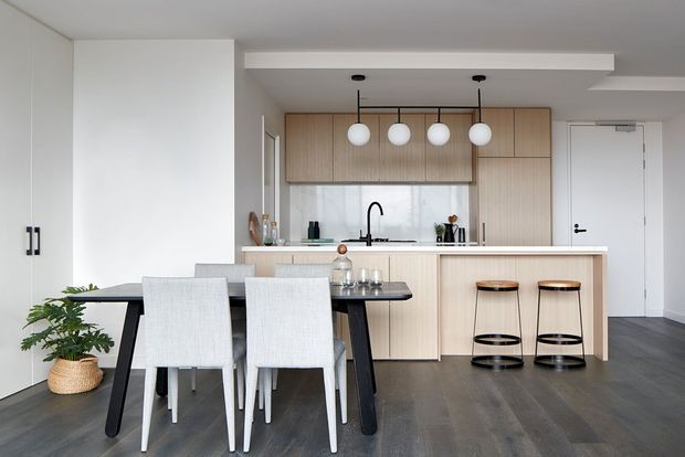 Bright contemporary open-plan kitchen and dining area with light timber cabinetry, a white island bench, black tapware and fittings, pendant globe lighting, and a dark dining table with upholstered chairs. Dark timber-look flooring contrasts with the pale cabinetry and walls.