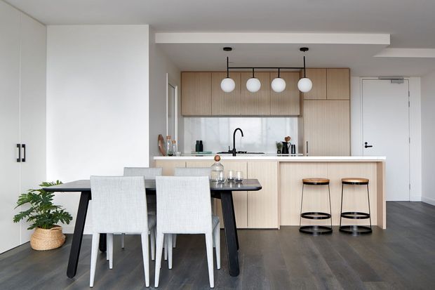 A bright, contemporary open-plan kitchen and dining area. The kitchen features full-height light timber cabinetry, a white benchtop and integrated island, black tapware and hardware, and four globe pendant lights. In the foreground is a dark dining table with upholstered dining chairs, while two black-framed stools sit at the island. Dark timber-look flooring and white walls create a clean, balanced palette.